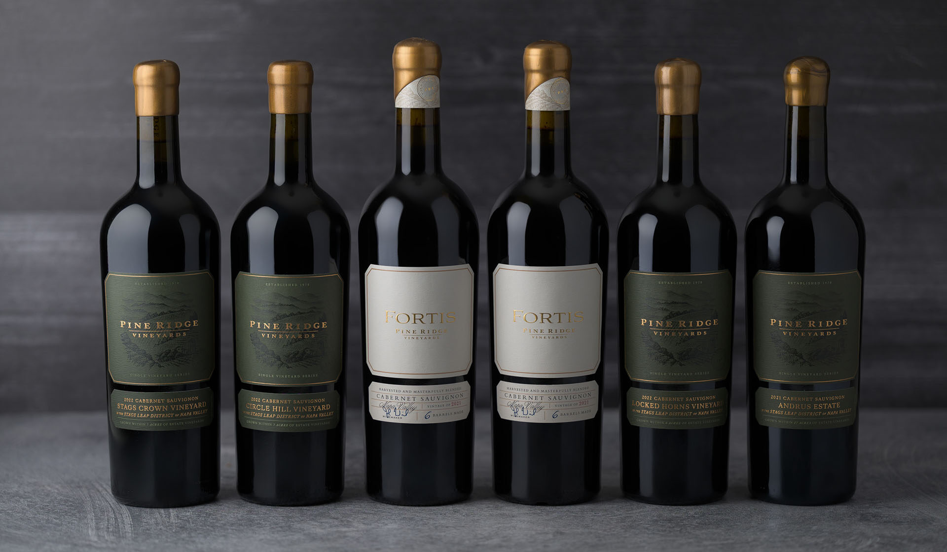 Six bottles of red wine are lined up against a dark background. Four bottles have dark green Pine Ridge Vineyards labels, while two in the center have cream-colored Fortis labels. All bottles have gold foil on the necks.