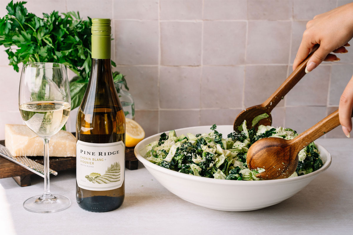 A person tosses a green salad in a white bowl with wooden utensils. Beside the bowl are a bottle of Pine Ridge wine, a glass of white wine, a wedge of cheese, a lemon, and fresh parsley on a light countertop.