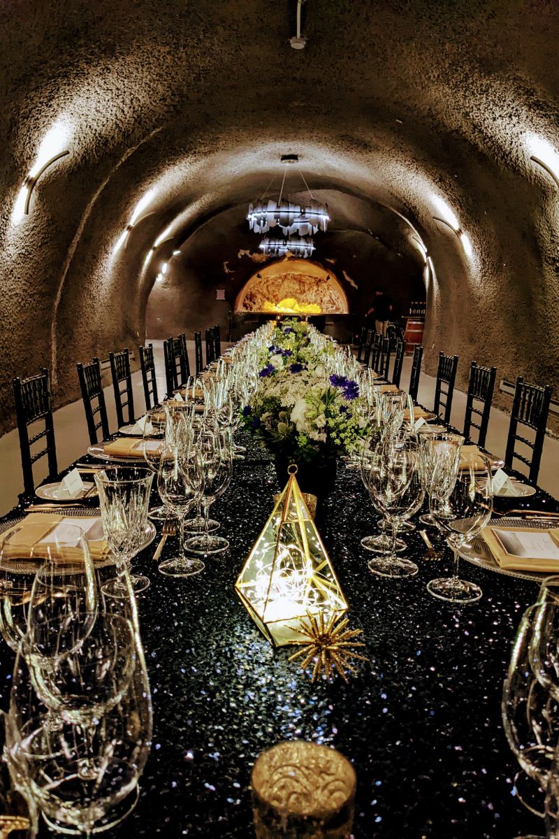 A long dining table in a tunnel-like room is set with glassware, plates, and floral centerpieces. A geometric lantern with lights is in the foreground. Black chairs line both sides of the table.