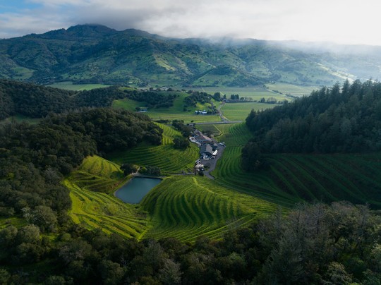 Aerial view of green hills with rows of terraced fields, a small body of water, and several buildings nestled in the valley, surrounded by forested areas and distant mountains under a cloudy sky.