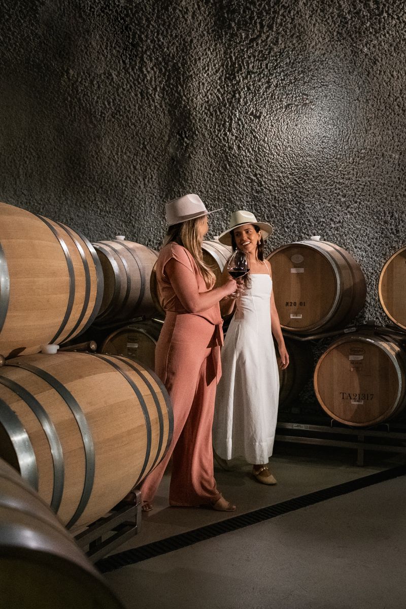Two women in hats stand and talk next to stacks of large wooden wine barrels in a dimly lit cellar. One woman wears a pink outfit, the other a white dress, and they appear to be sharing a glass of wine.
