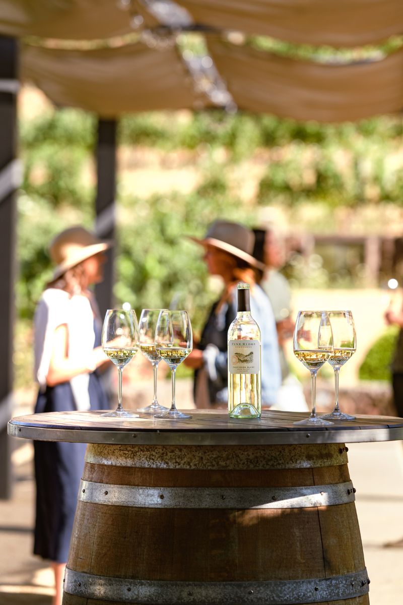 A wine bottle and four glasses sit on top of a wooden barrel. Two people wearing hats stand in the blurred background outdoors, talking under a canopy.