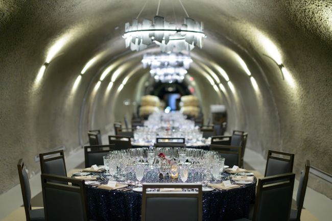 A long dining table is set with glassware, plates, and napkins inside a tunnel-like room with arched ceilings and modern chandeliers. Multiple chairs surround the table and wine barrels are visible in the background.