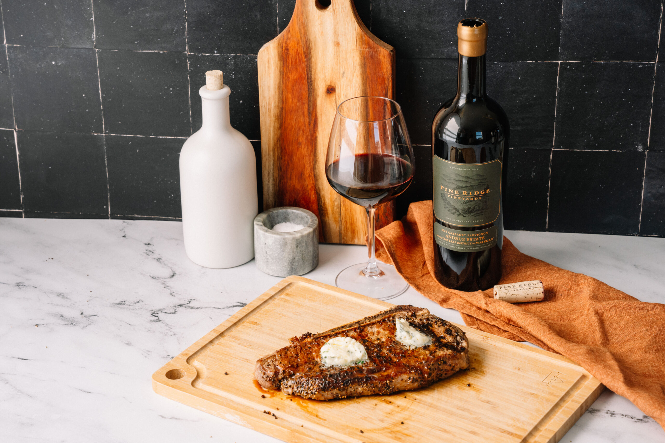 A cooked steak with a dollop of butter sits on a wooden cutting board next to a glass and a bottle of red wine, a small container of salt, a white ceramic bottle, and a wooden serving board on a marble countertop.