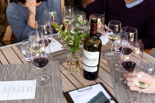 Four people sit around a wooden table set with wine glasses, a bottle of wine, menus, a small vase with greenery, and a clipboard. Some people are holding glasses or resting hands on the table.