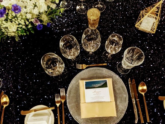 A formal table setting with gold utensils, a glass dinner plate, a menu card on a gold napkin, multiple wine glasses, a candle, a geometric ornament, and a floral centerpiece on a dark, glittery tablecloth.