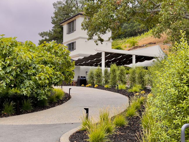 A curved driveway leads to a white two-story building with large windows, surrounded by green bushes, trees, and neatly landscaped plants. A covered patio area is visible on the right side of the building.