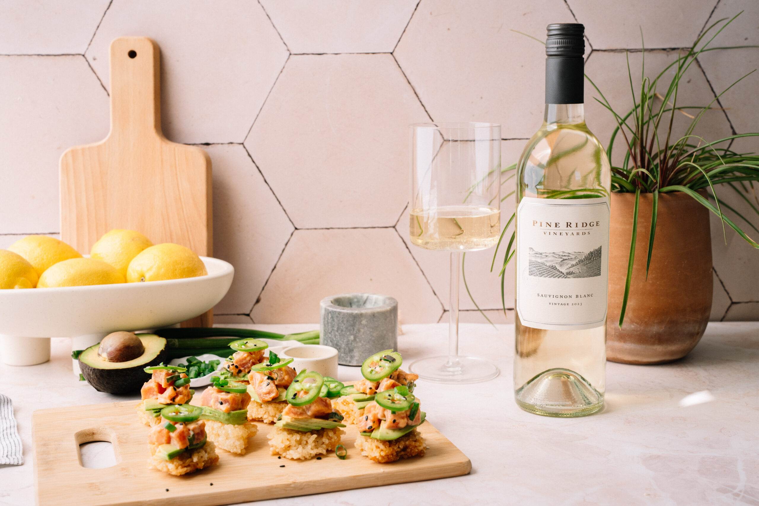 A bottle of white wine and two glasses are on a counter next to a plant, a bowl of lemons, an avocado, and a cutting board with sliced appetizers topped with vegetables and herbs.