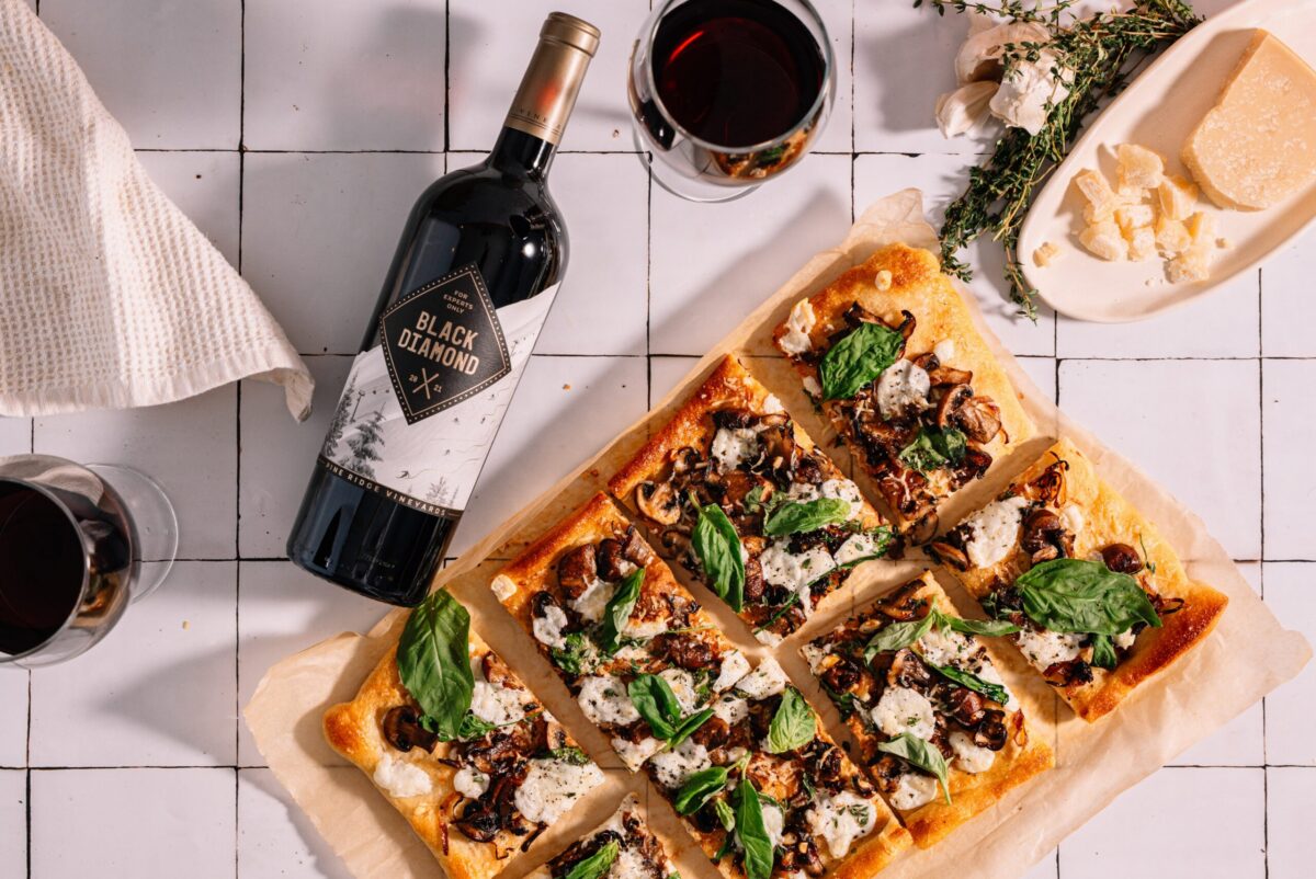 A sliced rectangular pizza with cheese, mushrooms, and basil on parchment paper sits on a tiled surface next to a bottle of Black Diamond wine, two filled wine glasses, and a plate with cheese, garlic, and herbs.