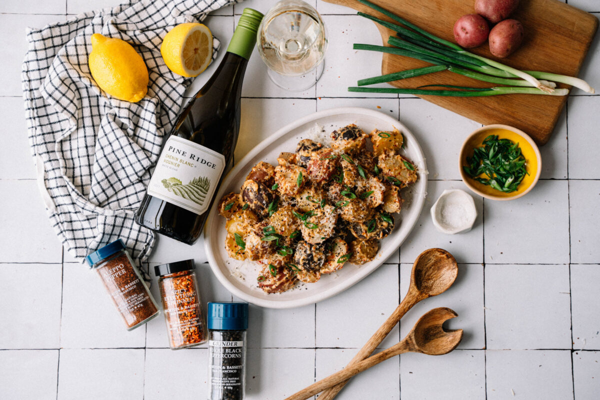 A white plate of seasoned roasted potatoes sits on a tiled surface with a bottle of white wine, a glass of wine, lemons, spices, wooden utensils, green onions, parsley in a bowl, and red potatoes on a cutting board.