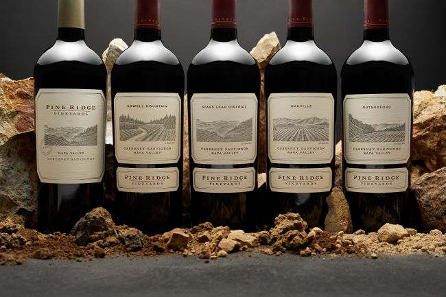 Five bottles of Pine Ridge Vineyards Cabernet Sauvignon stand upright in a row on soil and rocks, with detailed labels and a dark background. The bottles are evenly spaced and lit from the front.