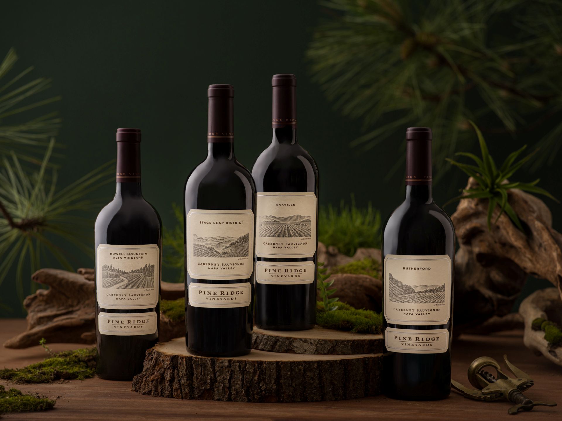 Four bottles of Pine Ridge Vineyards Cabernet Sauvignon are arranged on a table with moss, branches, pine needles, and a wine opener, against a dark green background.