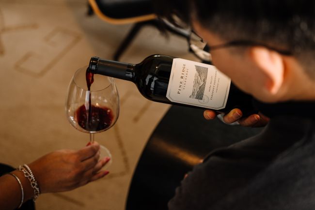 A person pours red wine from a bottle labeled PINE RIDGE into a glass held by another person with a bracelet. The scene takes place indoors on a carpeted floor.