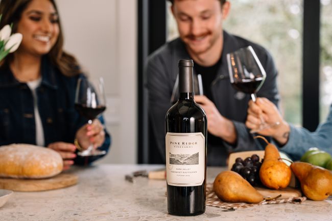 A bottle of Pine Ridge wine is placed on a kitchen counter with pears, grapes, a loaf of bread, and a green apple nearby. Two people holding wine glasses are sitting in the background, smiling at each other.
