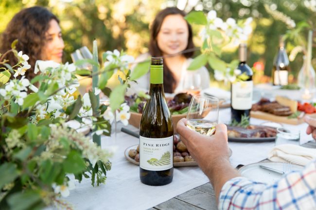 A group of people sit around an outdoor table set with food and flowers. A person holds a glass of white wine, with a bottle of Pine Ridge Chenin Blanc prominently displayed in the foreground.
