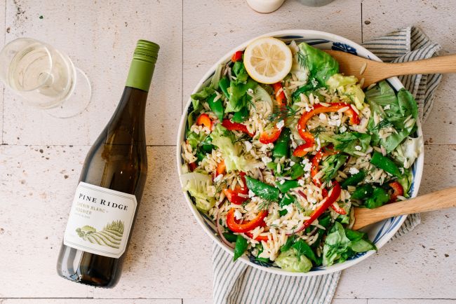 A bowl of salad with mixed greens, red bell peppers, orzo, and a lemon slice sits on a striped napkin next to a glass of white wine and a bottle labeled Pine Ridge. Wooden salad tongs are in the bowl.