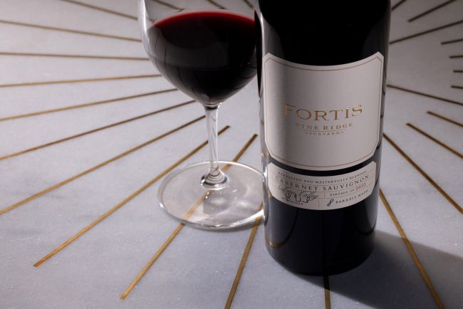 A bottle of Fortis Pine Ridge Cabernet Sauvignon stands next to a glass of red wine on a light gray surface with gold lines radiating outward.