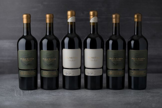 Six bottles of wine stand in a row against a gray background. The three on the left and the two on the right have green labels; the two bottles in the center have white labels. All bottles have gold caps.