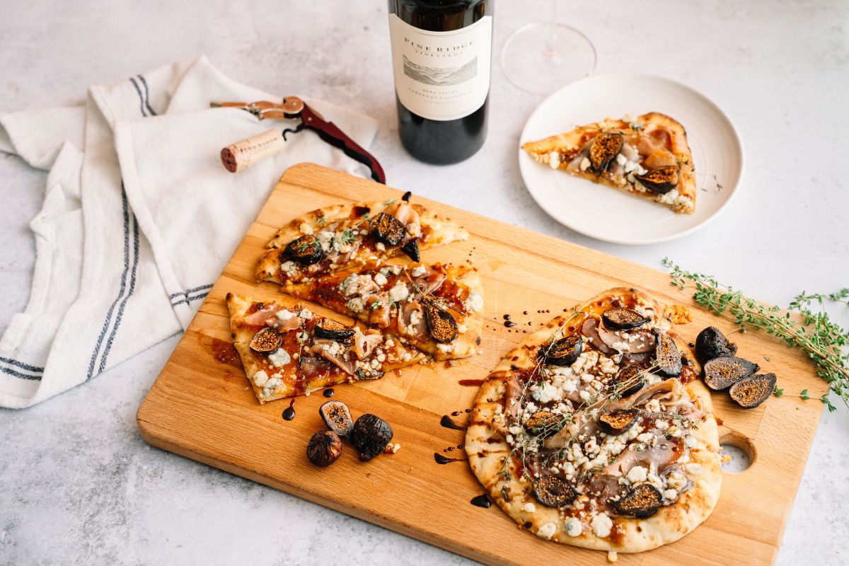 A wooden board with flatbread pizzas topped with figs, cheese, and herbs sits on a light surface. A wine bottle, glass, corkscrew, napkin, and fresh thyme are nearby.