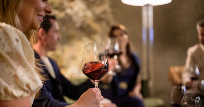 Four people sit together indoors, each holding a glass of red wine. The focus is on the woman in the foreground, smiling and lifting her glass. The background is softly blurred, showing a warm, relaxed atmosphere.