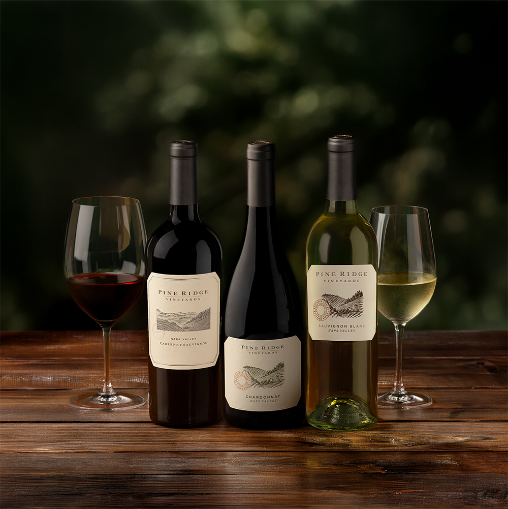 The Curated Trio: Three bottles of Pine Ridge wine—one red, one white, and one Chardonnay—stand on a wooden table, each beside a filled glass, with a green, blurred background.