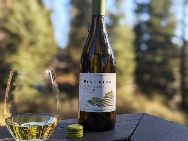 A bottle of Pine Ridge Chenin Blanc + Viognier white wine stands on a wooden surface next to a filled wine glass and a yellow bottle cap, with trees and greenery blurred in the background.