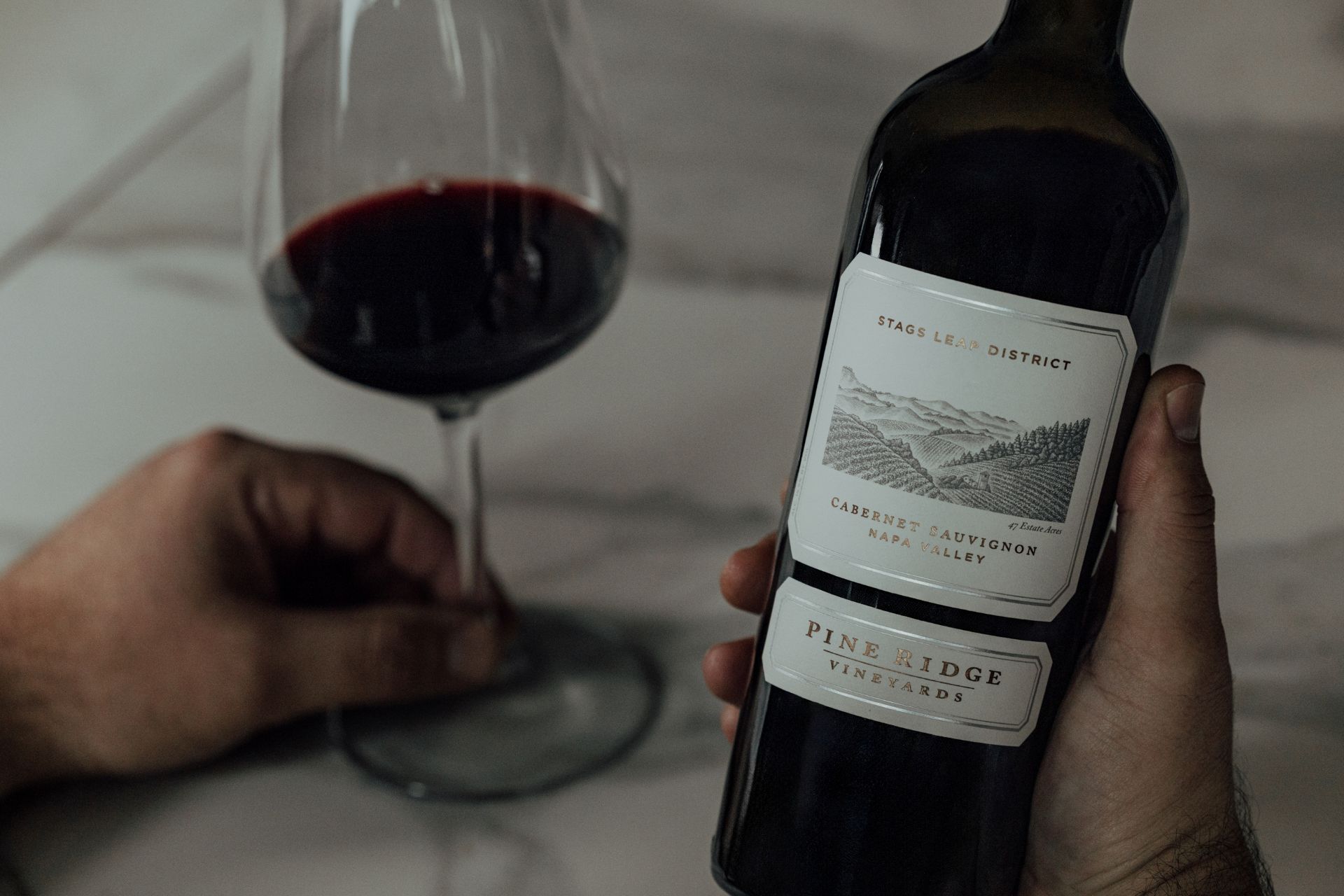 A person holds a glass of red wine in one hand and a bottle of Pine Ridge Vineyards Cabernet Sauvignon in the other, on a white marble surface.