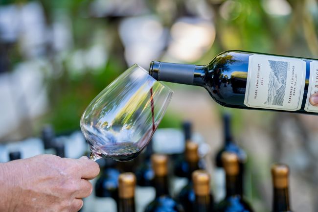 A person pours red wine from a bottle into a glass outdoors. Several wine bottles with gold-colored caps are visible in the background.
