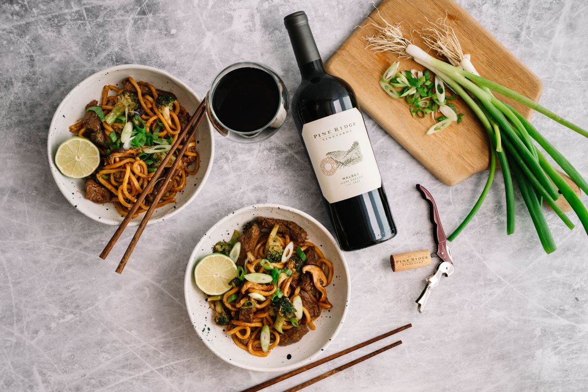 Two bowls of noodle stir-fry with lime slices and chopsticks are placed next to a glass and bottle of red wine. A cutting board with chopped green onions, a cork, and a corkscrew are nearby on a gray surface.