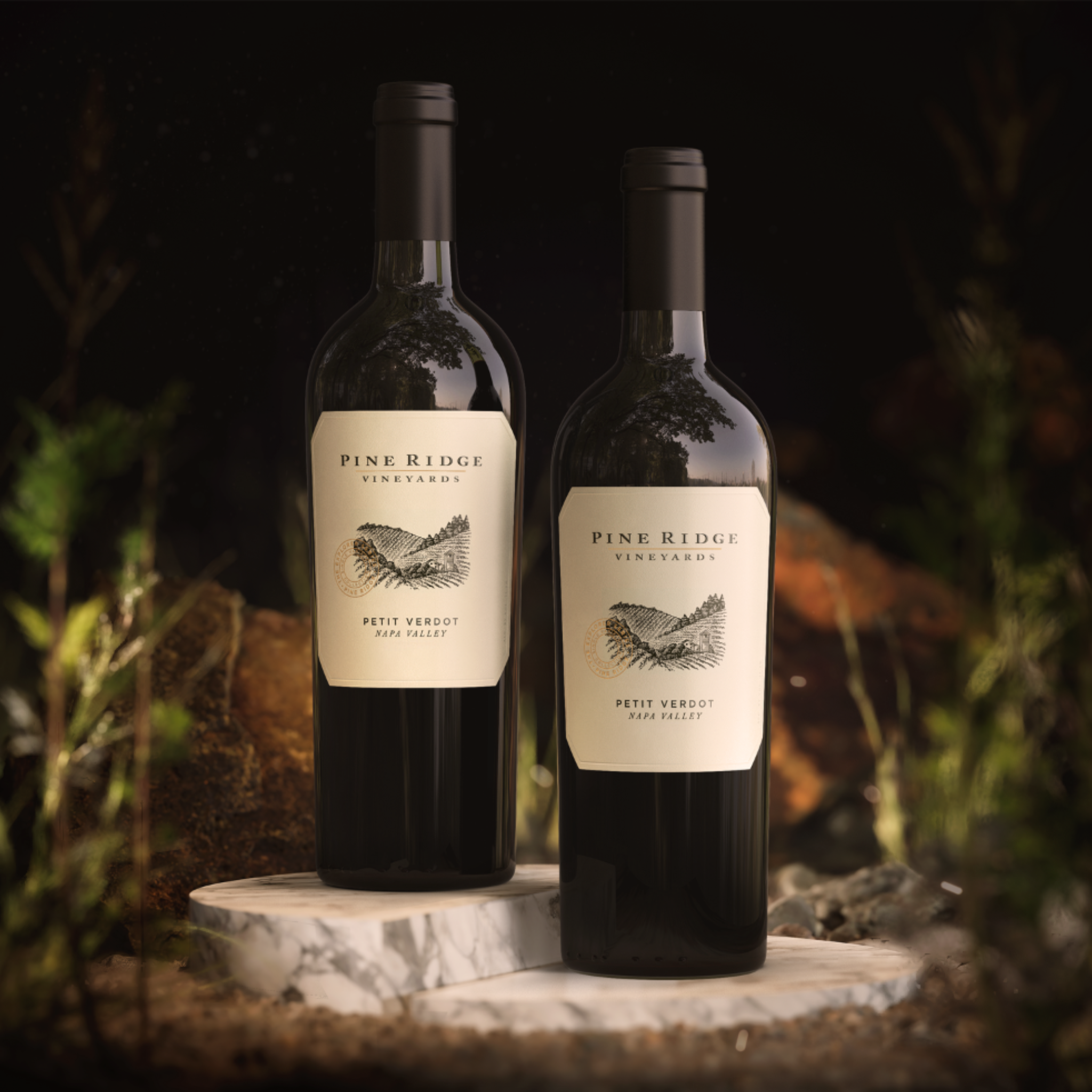 Two bottles of Pine Ridge Vineyards Petit Verdot wine are displayed on marble slabs outdoors, with lush foliage and rocks in the background. Both bottles feature cream-colored labels with elegant vineyard illustrations, highlighting the Petit Verdot varietal.