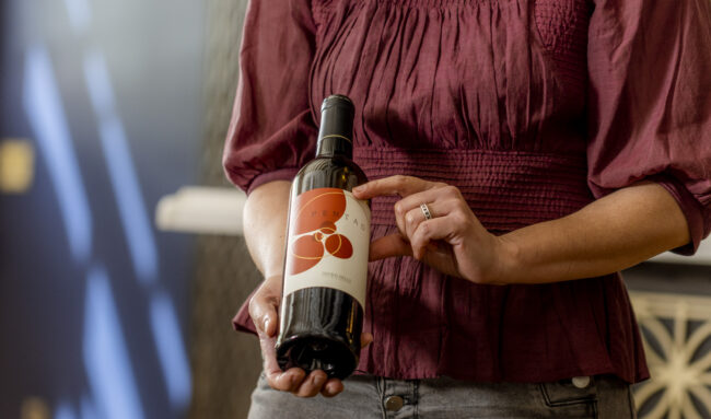 A person wearing a maroon top holds a bottle of wine with a white label featuring an orange graphic and text. The person points at the label with one finger.