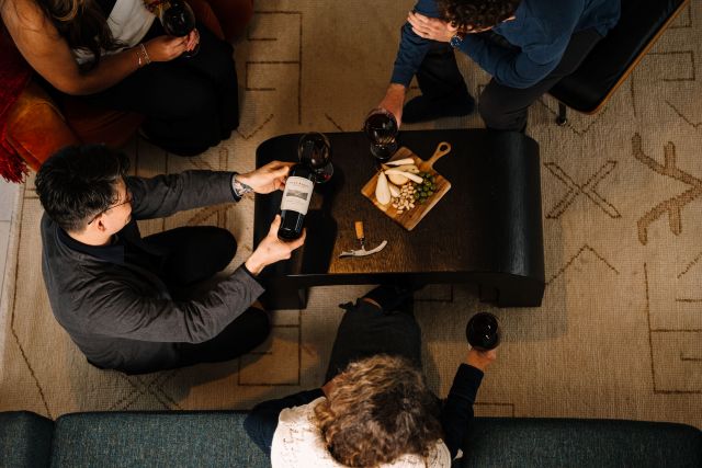 Four people sit around a dark coffee table with wine glasses, a bottle of wine, a corkscrew, and a wooden board with cheese and snacks on a beige patterned rug. The scene is viewed from above.