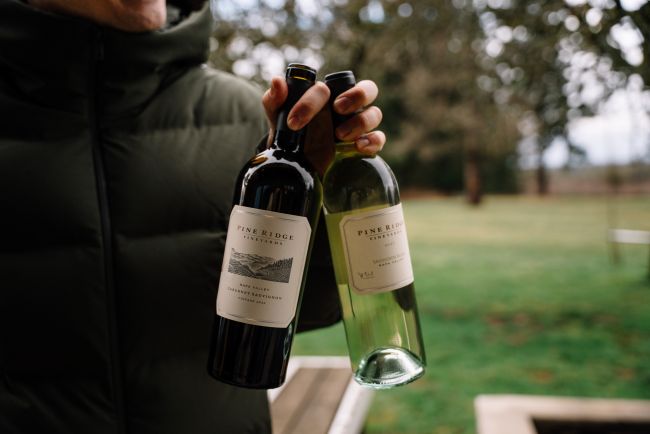 A person wearing a dark jacket holds two bottles of Pine Ridge wine, one red and one white, outdoors with a grassy area and trees in the background.