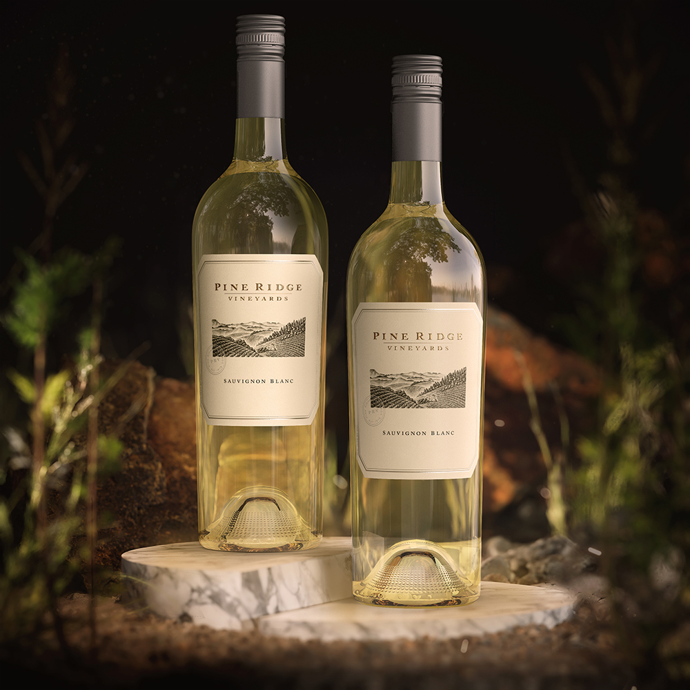Two bottles of Pine Ridge Vineyards Sauvignon Blanc stand on a marble slab, light yellow wine gleaming in each. Greenery and rocks are softly blurred in the background, highlighting the elegance of this Sauvignon Blanc.