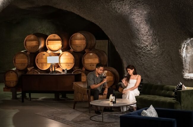 A man and woman sit on a green couch inside a wine cellar with stacked wooden barrels. They are talking and holding glasses of wine. A table with a wine bottle and two glasses is in front of them.