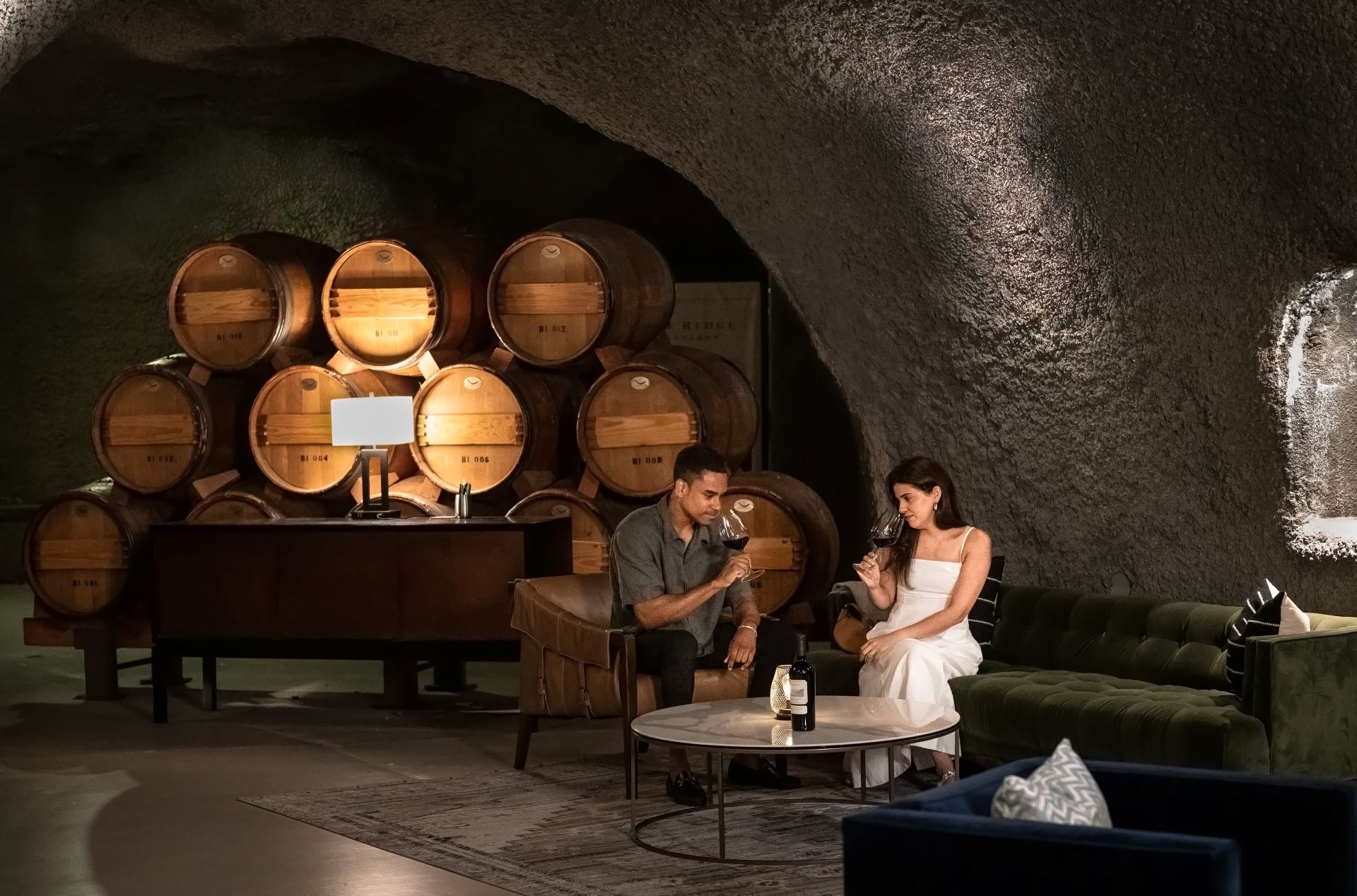 A man and woman sit on a green couch inside a wine cellar with stacked wooden barrels. They are talking and holding glasses of wine. A table with a wine bottle and two glasses is in front of them.