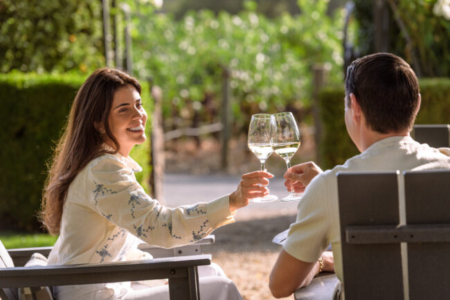 Two people sit outdoors in wooden chairs, holding glasses of white wine and making a toast, with a vineyard visible in the background.