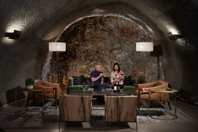 Two people sit on a green sofa in a cave-like room with stone walls, tasting wine. They are flanked by modern lamps and surrounded by contemporary furniture, including leather chairs, a coffee table, and a potted plant.