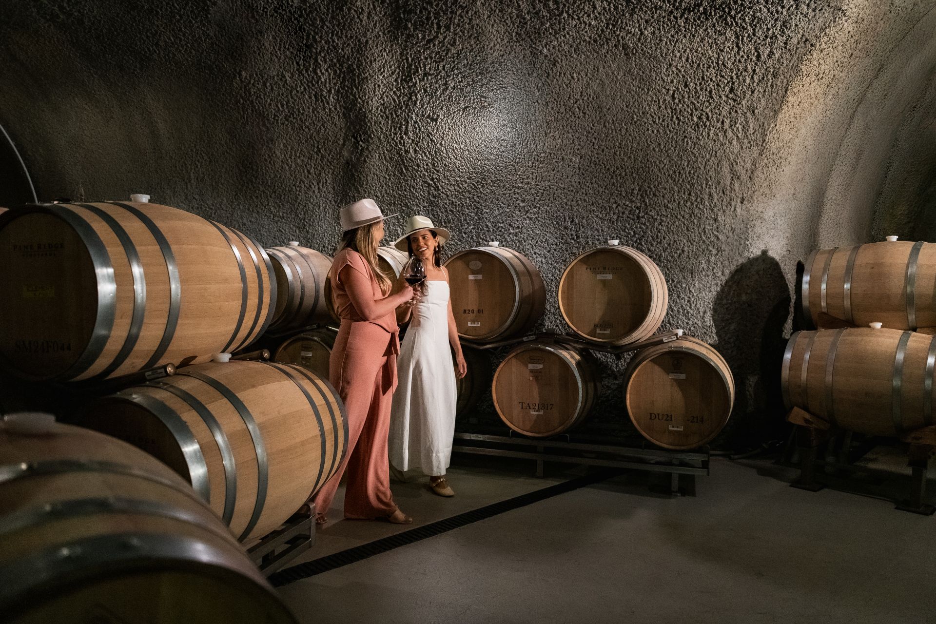 Two women in hats stand and talk inside a wine cellar, surrounded by large wooden barrels placed on metal racks against textured stone walls.
