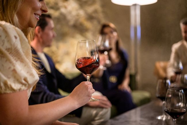 Four people sit together indoors, each holding a glass of red wine. The focus is on the glass in the foreground. The background is softly blurred, showing relaxed conversation in a cozy, warmly lit setting.