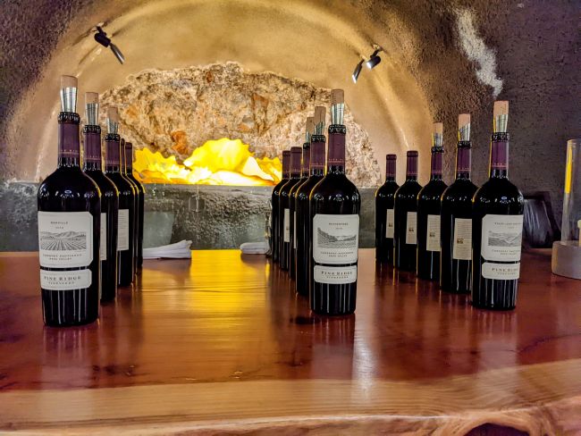 Twenty bottles of red wine are arranged in two parallel rows on a wooden table in front of a stone oven with a glowing yellow light inside. The scene has a rustic, cellar-like atmosphere.