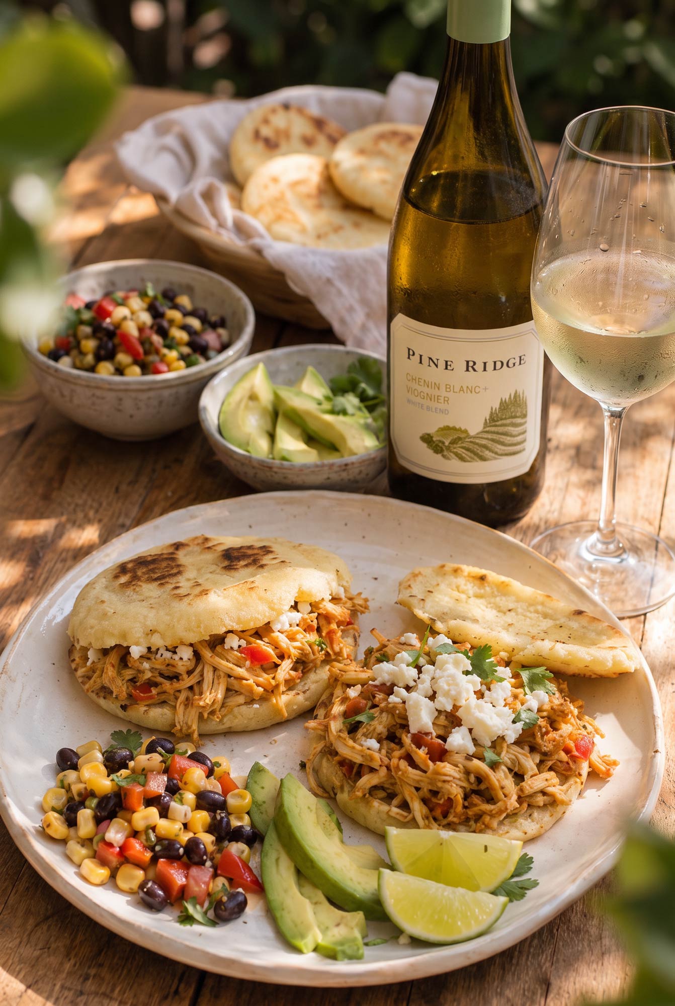 A plate with two arepas filled with shredded chicken, sliced avocado, and cheese sits next to a glass and bottle of white wine. Nearby are bowls of corn salad, avocado slices, and arepas in a basket.