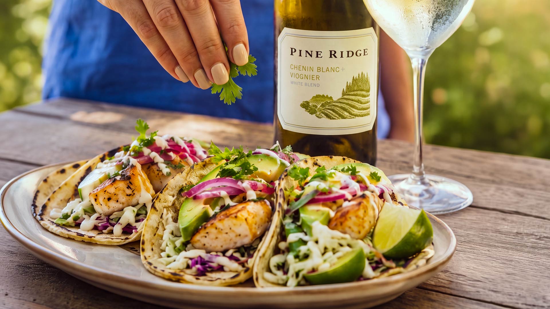 A hand places cilantro on fish tacos served with lime wedges on a plate. A bottle of white wine labeled Pine Ridge and a glass of wine are on a wooden table outdoors.