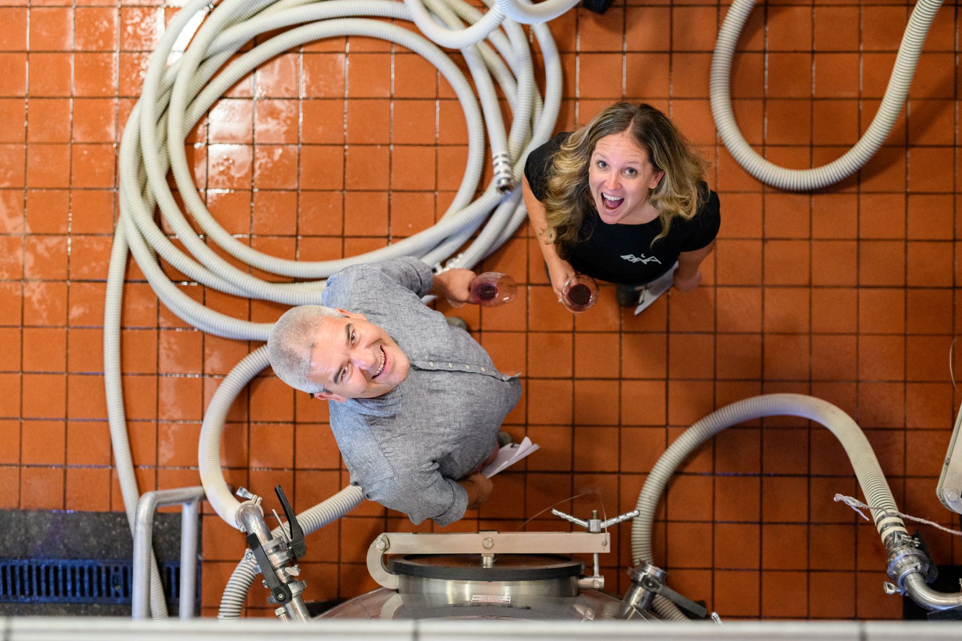 Two people stand on a red tiled floor, looking up at the camera, surrounded by large hoses and metal brewing equipment. Both are holding drinks and smiling.