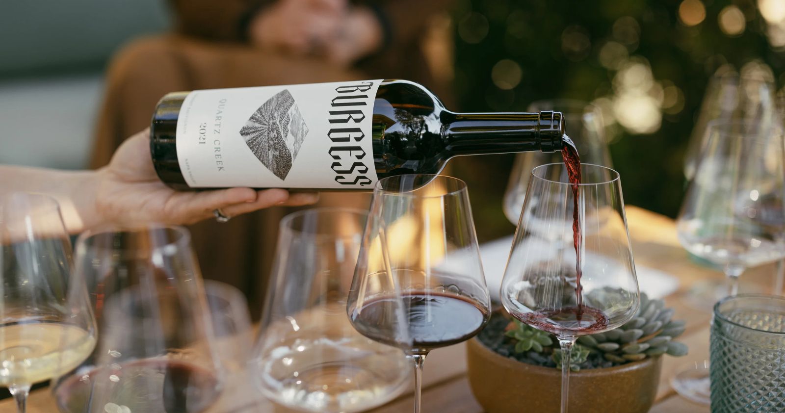 A hand pours red wine from a Burgess bottle into a glass on a wooden table set with several wine glasses, a succulent plant, and a blurred background.