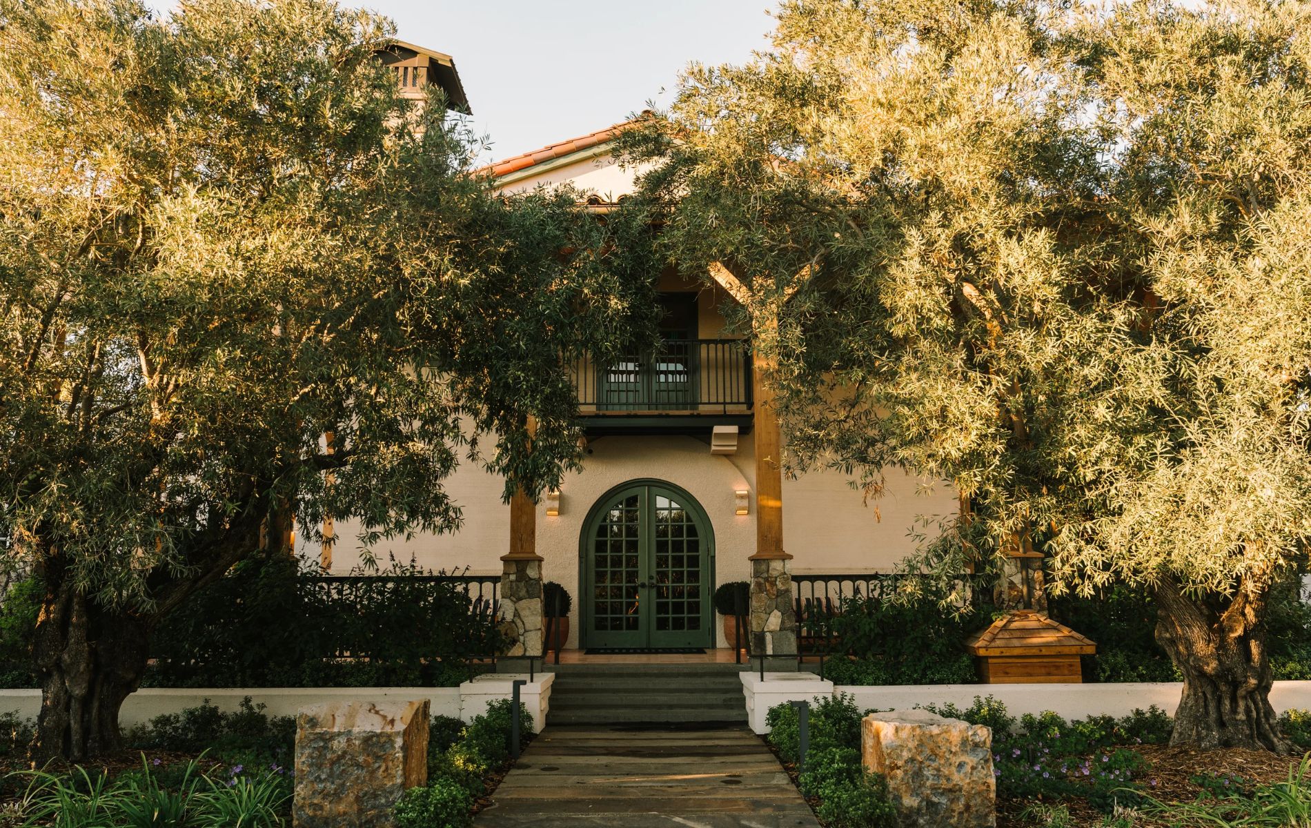 A two-story house with a green arched door and a small balcony, partially obscured by large leafy trees. Stone steps and a path lead to the entrance, with landscaped plants on either side.