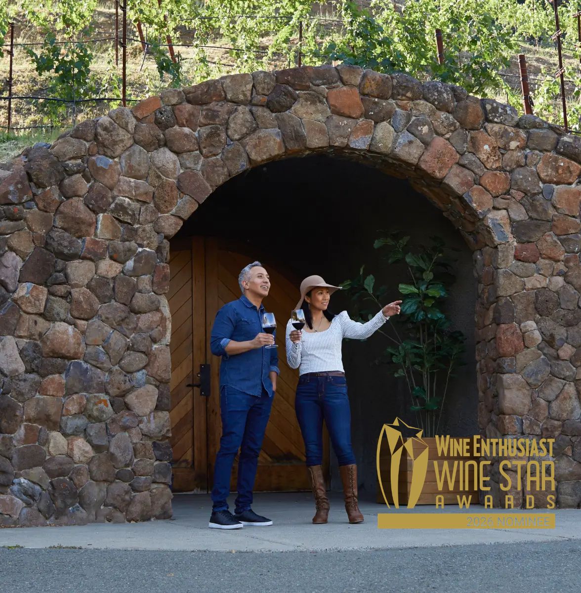 Two people stand in front of a stone archway holding wine glasses. The woman points to something off-camera. Behind them, large wooden doors are visible. The Wine Enthusiast Wine Star Awards 2023 logo overlaps the lower right corner.